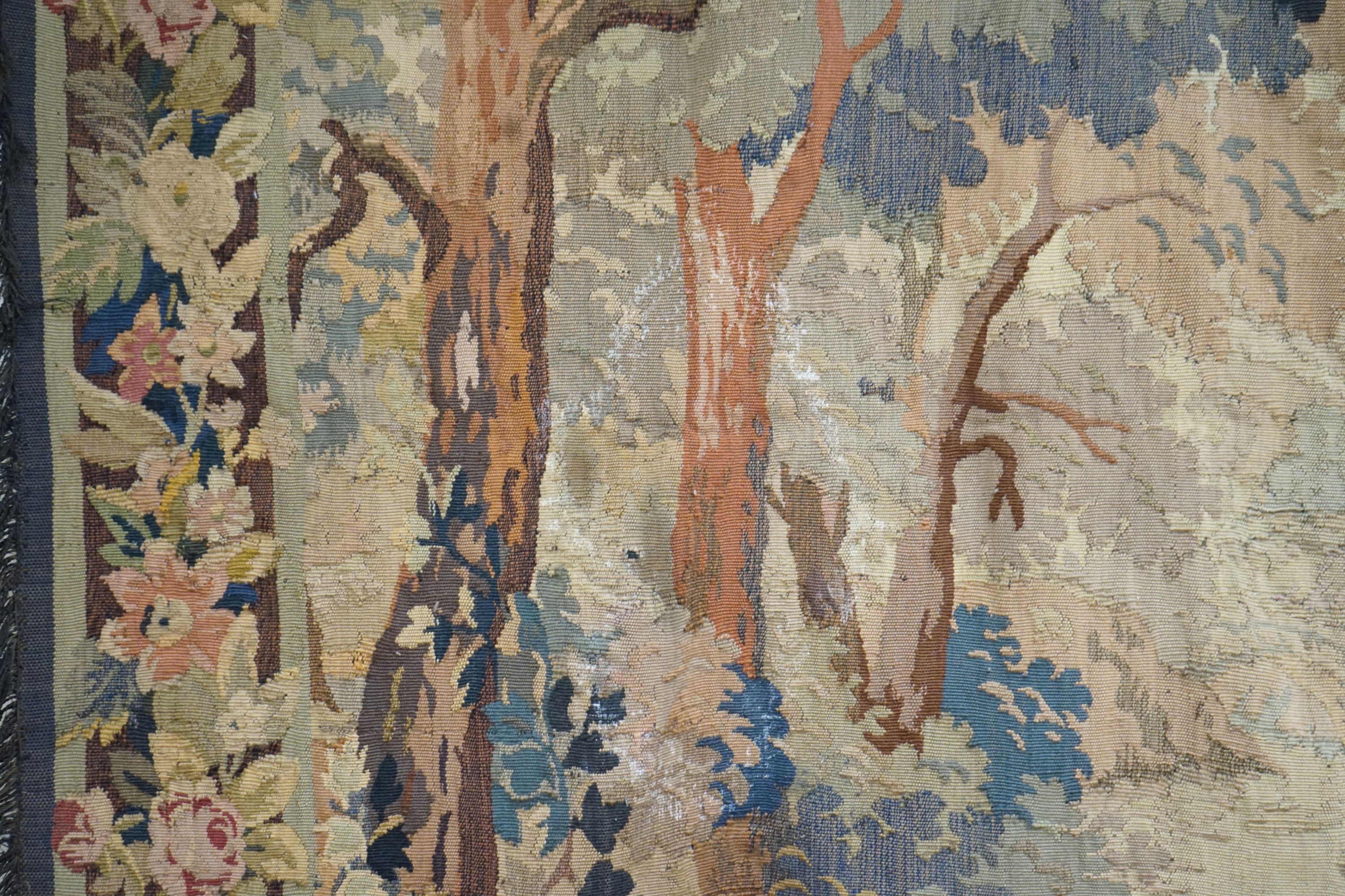 A late 19th / early 20th century French tapestry depicting a woodland scene with trees and flowers with a floral border woven in autumnal colours